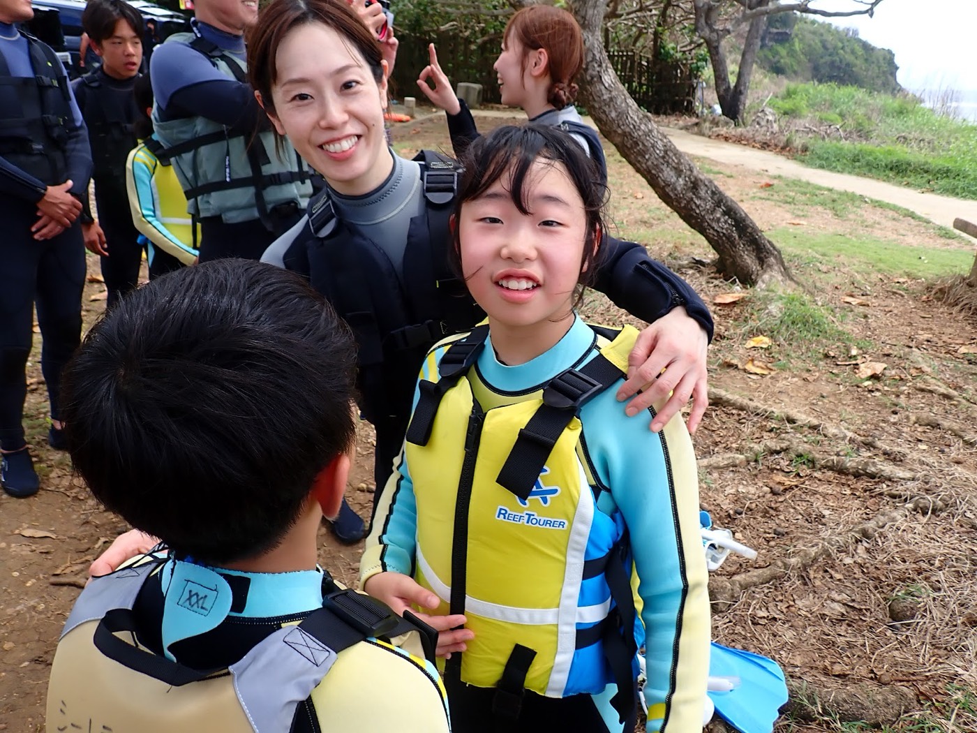 ライフジャケットを着用した子供と笑顔の保護者がいる陸上の風景。天然水族感が広がる