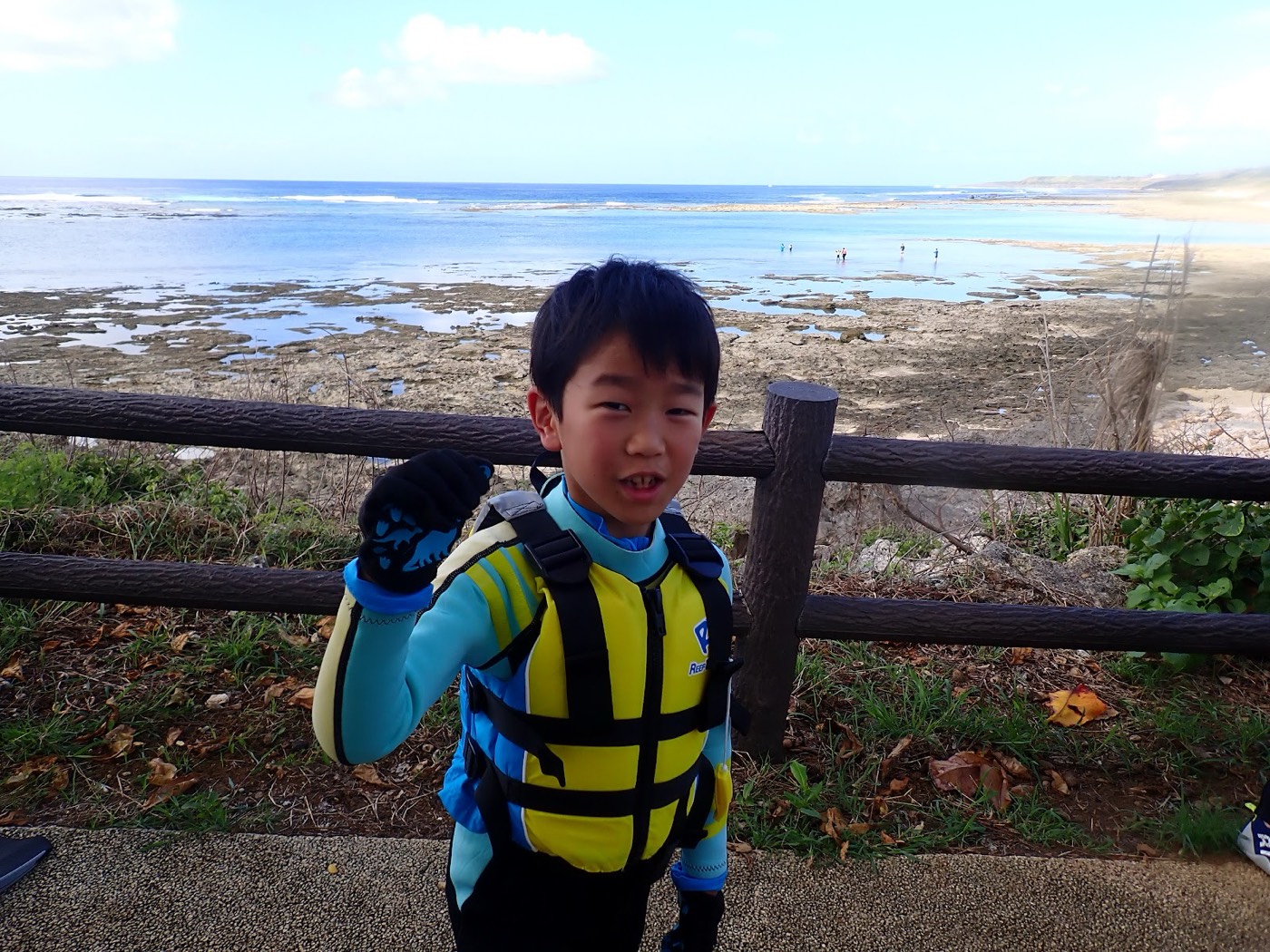 ライフジャケットを着用した子供が、準備中の器材と広い干潟、青空を背景にポーズ。天然水族感
