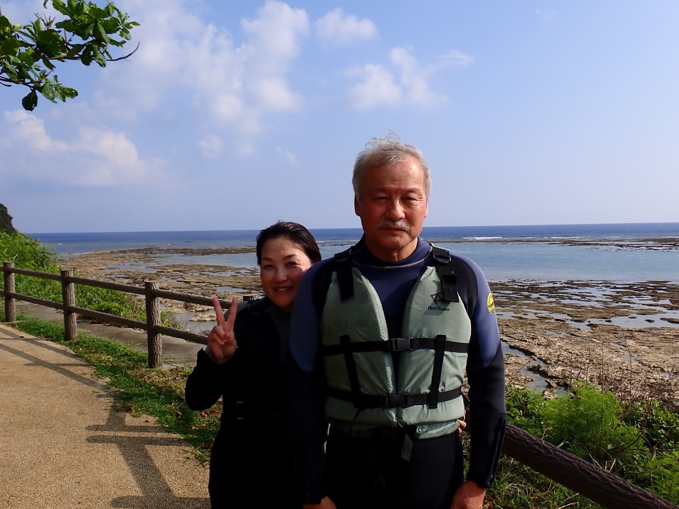天然水族感の海岸のリーフと青い空を背景にピースサインで立つ参加者。天然水族館での自然体験シュノーケル